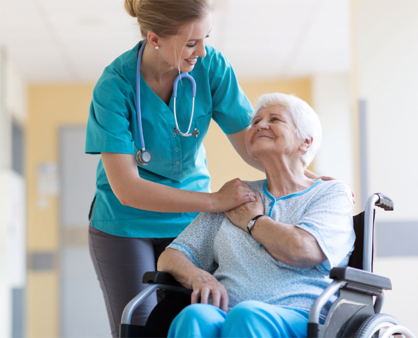 An elderly woman in a wheelchair smiles and clasps hands with a medical caretaker