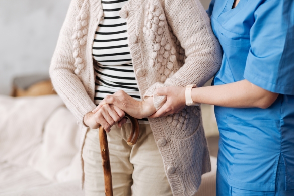 an elder stands, supported by a wooden cane and a medical professional wearing blue scrubs